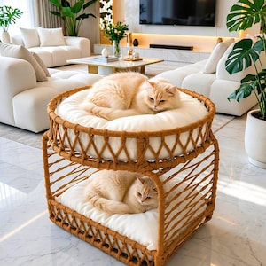 Puede incluir: Una cama para mascotas de dos niveles construida con ratán marrón tejido, con una base rectangular y una parte superior circular. Cada nivel cuenta con un cojín blanco grueso y acolchado. Dos gatos de pelo largo color crema descansan en ella.