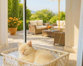 May include: A cream-colored cat resting in a rectangular, off-white macrame window perch. The perch features a woven rope frame with a soft white cushion inside, suspended against a glass window by two thin cables attached with suction cups.