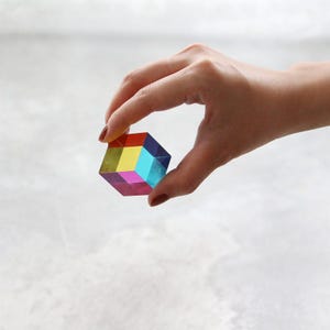Puede incluir: Un cubo geométrico multicolor con facetas rojas, amarillas, rosas y azules. El cubo se sostiene entre los dedos sobre un fondo neutro. El diseño del cubo es una versión moderna de un rompecabezas clásico.