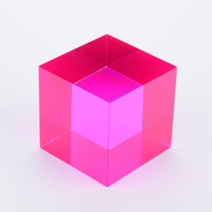 May include: A pink acrylic cube with a smaller pink cube inside. The larger cube is translucent, and the smaller cube is opaque.