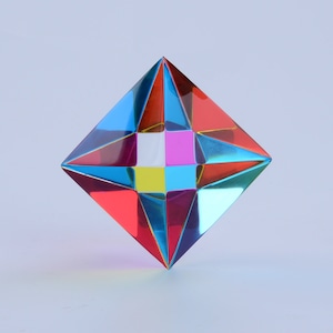 Puede incluir: Una escultura de cristal geométrico multifacético, iridiscente, con un diseño en forma de estrella. El cristal está formado por múltiples facetas triangulares en tonos de rojo, azul, amarillo, rosa y blanco.