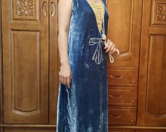 May include: A blue velvet sleeveless dress with a gold lining and a decorative bow design. The dress has a V-neck and a flowing skirt. A pearl necklace is draped over the dress. The dress is a vintage style.