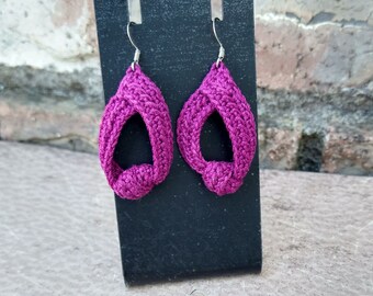 May include: A pair of pink crocheted earrings with a knotted design. The earrings are hanging on a black display stand.