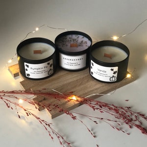 May include: Three black metal container candles with wooden wicks sit on a wooden board. The candles are labeled "Pumpkin Pie", "Entspannung", and "Vanille". The "Pumpkin Pie" candle has a white wax, the "Entspannung" candle has a lavender wax, and the "Vanille" candle has a white wax.