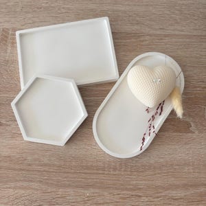 May include: Three white decorative trays in different shapes: a rectangle, a hexagon, and an oval. The oval tray holds a heart-shaped candle with a textured surface, and dried floral accents. The trays are arranged on a wooden surface.