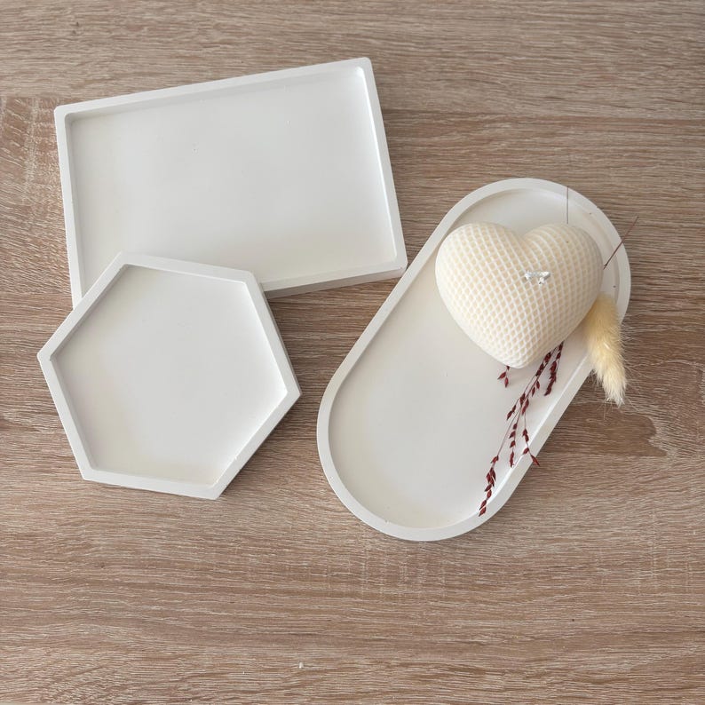 May include: Three white decorative trays in different shapes: a rectangle, a hexagon, and an oval. The oval tray holds a heart-shaped candle with a textured surface, and dried floral accents. The trays are arranged on a wooden surface.
