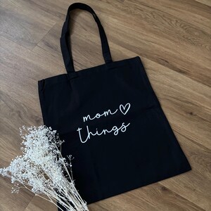 Könnte beinhalten: Schwarze Tragetasche mit den Worten "mom things" in weißer Schrift und einem Herzsymbol. Die Tasche hat lange schwarze Henkel und liegt auf einer Holzoberfläche. Ein Bund weißer Trockenblumen liegt neben der Tasche.