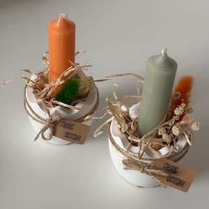 Könnte beinhalten: Zwei weiße, eiförmige Kerzenhalter mit orangefarbenen und grünen Kerzen. Jeder Halter ist mit getrocknetem Gras, dekorativen Zweigen und einem kleinen Etikett mit der Aufschrift "Frohe Ostern" verziert. Die Halter sind mit Bindfaden gebunden.