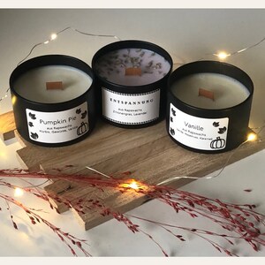 May include: Three black metal candles with wooden wicks sit on a wooden board. The candles are labeled "Pumpkin Pie", "Entspannung", and "Vanille". Each candle has a white label with text and a pumpkin graphic. The candles are surrounded by decorative lights.