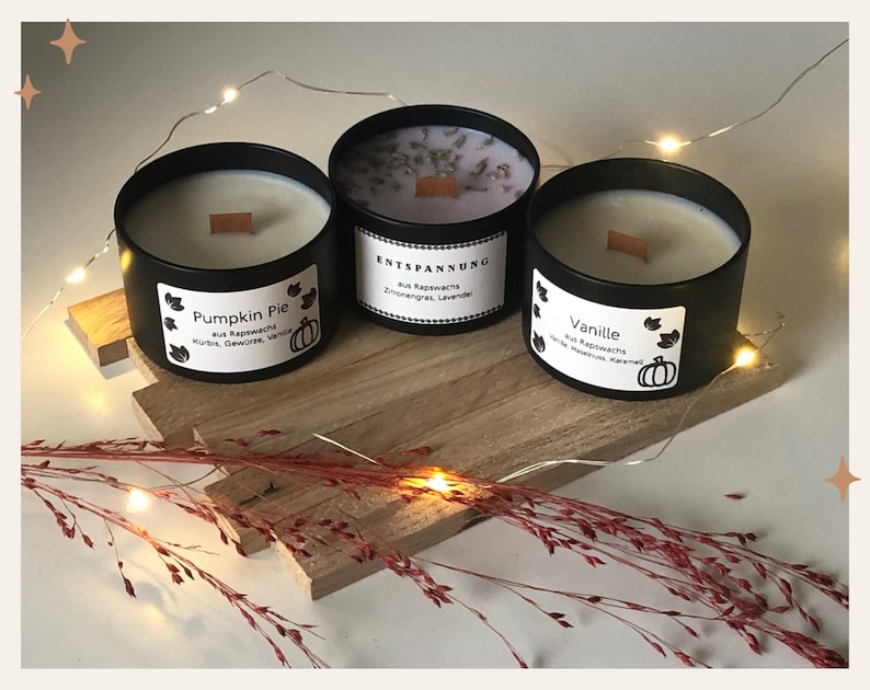 May include: Three black metal candles with wooden wicks sit on a wooden board. The candles are labeled "Pumpkin Pie", "Entspannung", and "Vanille". Each candle has a white label with text and a pumpkin graphic. The candles are surrounded by decorative lights.