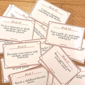 May include: A collection of cream-colored cards with brown borders and text. The cards are scattered on a wooden surface. Each card features a different activity suggestion, such as "Visit a park" or "Decorate your mirror."