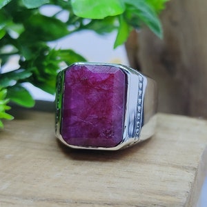 Puede incluir: Un anillo de plata con una gran piedra preciosa de rubí rojo oscuro, rectangular y facetada, engastada en el centro. La banda de metal tiene un acabado pulido con una textura decorativa de cuentas a los lados del engaste de la piedra.