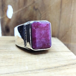 Può includere: Un anello in argento con una grande pietra preziosa rubino rosa intenso, rettangolare e sfaccettata, incastonata in una fascia di metallo lucido. Il lato della fascia presenta una fila verticale di piccole rientranze circolari lungo il bordo.