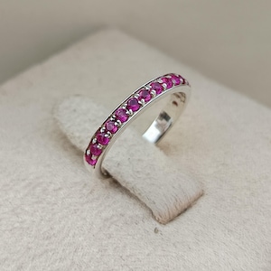 May include: A silver band ring featuring a row of small, round, vibrant pink gemstones set in a prong setting along the top half of the band. The metal has a polished, reflective finish, and the stones are uniform in size and colour.
