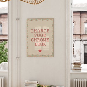 May include: A framed print with the text "CHARGE YOUR CHROME BOOK" in red cross-stitch style. The print has a beige background with a red heart at the bottom and a decorative border with green, red, and orange floral patterns. The print is hanging on a white wall.