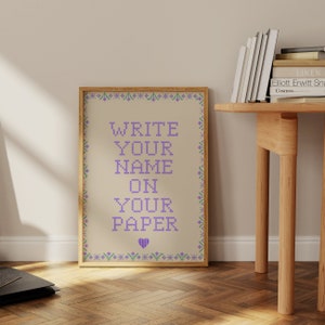 Può includere: Una stampa incorniciata con uno sfondo beige e un design a punto croce viola che dice "WRITE YOUR NAME ON YOUR PAPER". La stampa è circondata da una cornice dorata.