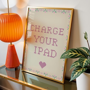 May include: A framed cross-stitch art piece featuring the text "CHARGE YOUR IPAD" in purple, surrounded by a pink and green floral border with a small purple heart at the bottom, all set against a cream-colored background within a gold frame.