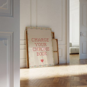 May include: A framed artwork with the text "CHARGE YOUR CHROME BOOK" in red cross-stitch style. The artwork has a cream background, a decorative border, and a red heart. It leans against a wall with other framed pieces.