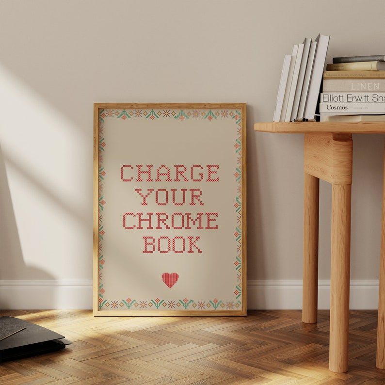 May include: A framed print with the text "CHARGE YOUR CHROME BOOK" in red cross-stitch style. The print has a beige background, a decorative border, and a red heart at the bottom. The frame is a light wood color.