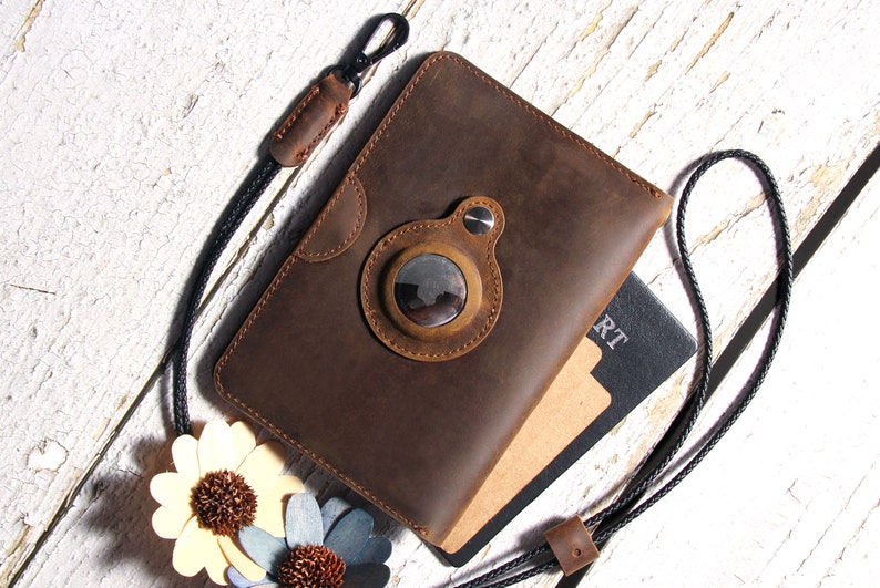 May include: A brown leather passport holder with a black strap and a round, metal accent. The holder is lying on a white wooden surface with a passport and two decorative flowers.