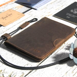May include: A brown leather passport holder with a black cord and a clasp. The holder has a subtle embossed design and the word "PASS" is visible on the front.