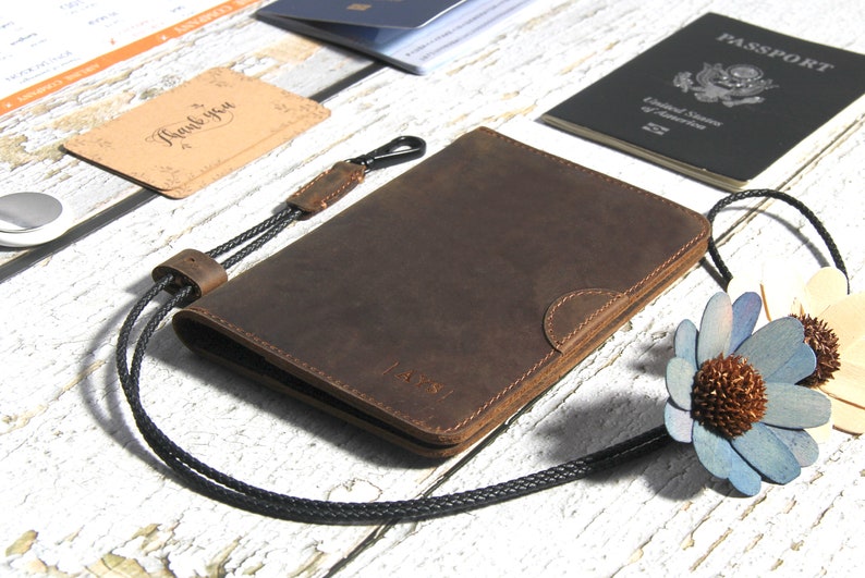 May include: A brown leather passport holder with a black cord and a clasp. The holder has a subtle embossed design and the word "PASS" is visible on the front.