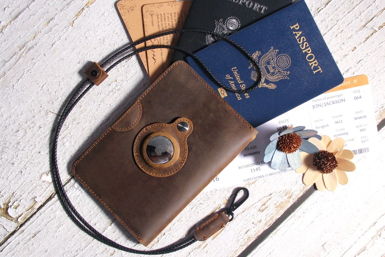 May include: A brown leather passport holder with a circular window and a black strap. The holder is lying on a white wooden surface with a passport, boarding pass, and two decorative flowers.