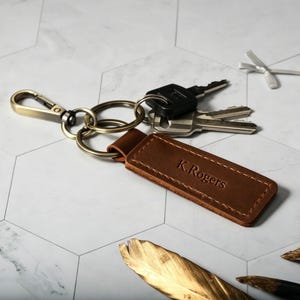 May include: A rectangular brown leather keychain featuring the embossed name K.Rogers. The leather tag is attached to a brass-toned metal split ring and a lobster clasp, which holds a set of three silver and black metal keys.