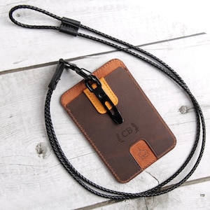 May include: A brown leather card holder with a tan accent tab and a black braided lanyard. The card holder features the embossed letters CB, a small leaf design, and a circular icon at the bottom. A black metal clip attaches the lanyard to the holder.