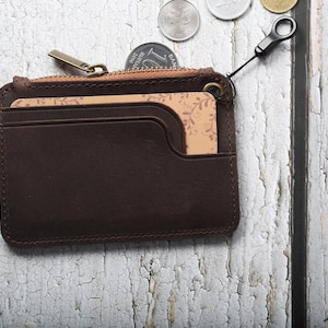Könnte beinhalten: Ein dunkelbraunes Leder-Kartenetui mit einem Reißverschlussfach oben und einer Vordertasche, die eine beige Karte mit zartem Blattmuster enthält. Das Etui verfügt über einen Metall-Reißverschlussanhänger und eine kleine schwarze Schlaufe.