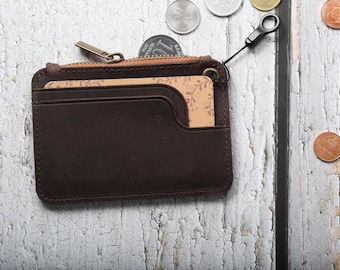 May include: A dark brown leather card holder with a zippered top compartment and a front pocket containing a tan card with a delicate leaf pattern. The wallet features a metal zipper pull and a small black loop attachment on the top right corner.