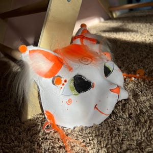 May include: A white cat mask with orange accents, including ears, whiskers, and a small party hat. The mask features mesh eye coverings, a smiling mouth, and is designed for costume or cosplay use.