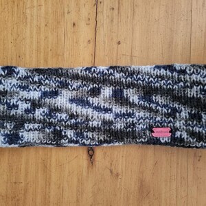 Könnte beinhalten: Gestricktes Stirnband in Blau-, Grau- und Weißtönen mit einem kleinen rosa Etikett. Das Stirnband besteht aus dickem, strukturiertem Garn, perfekt, um die Ohren warm zu halten. Das Accessoire ist für kaltes Wetter konzipiert.