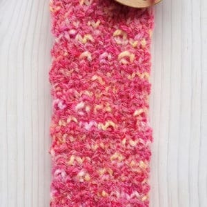 Könnte beinhalten: Ein handgestrickter Socken aus meliertem rosa und gelbem Garn. Ein rundes Pappetikett ist mit rosa Garn befestigt und zeigt eine Schleifenillustration sowie den Text "www.lykken.com", "HANDMADE" und "0-6 Monate" in schwarzer Tinte.