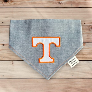May include: A gray denim dog bandana with a white embroidered letter "T" outlined in orange. The bandana has a small white tag that says "Paw Chic Threads handmade".