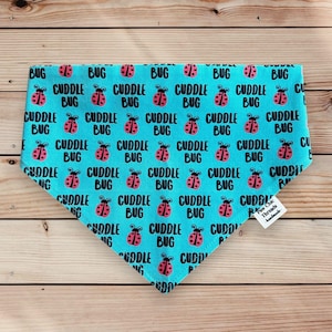 Puede incluir: Un bandana azul turquesa con un patrón repetido de mariquitas rojas y el texto "Cuddle Bug". El bandana tiene una etiqueta blanca con el texto "Two Chic Threads Handmade".
