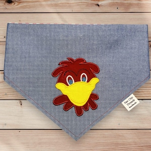 May include: A denim blue dog bandana with a cartoon bird applique. The bird has a yellow beak, blue eyes, and red feathers. The bandana is triangular and has a red stitched border. A small tag reads "Paw Chic Threads handmade."