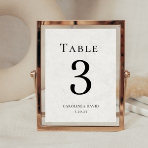 May include: A gold-framed table number card with a white marble background. The number "3" is printed in black, along with the text "TABLE" and "CAROLINE & DAVID 5.20.23".