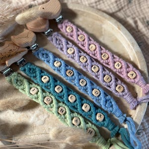 Könnte beinhalten: Eine Sammlung von Schnullerhaltern aus Makramee in Pastellfarben wie Grün, Blau und Lila. Jeder Halter hat Holzperlen mit Namen wie "ELENA", "MARIE", "FELIX" und "JONAH". Die Halter haben Vogel-förmige Holzaufsätze.