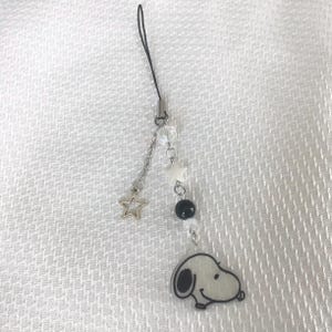 May include: A white and black charm with a cartoon dog face hangs from a black string with silver beads and stars.