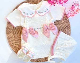 May include: A cream-colored knitted short-sleeve romper featuring a scalloped collar with pink and green floral embroidery, two large pink satin bows at the waist, matching knitted booties with floral trim, and a cable-knit bonnet with pink trim.