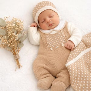 May include: A sleeping baby wearing a beige knit outfit with white polka dots, a matching hat, and a white collared shirt. A beige blanket with white polka dots is next to the baby. A small bouquet of dried flowers is on the left.