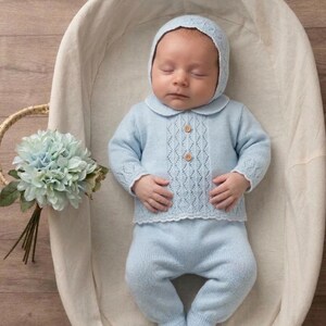 Könnte beinhalten: Ein hellblaues, gestricktes Baby-Outfit bestehend aus einem langärmeligen Pullover mit zartem Rautenmuster, zwei kleinen Holzknöpfen, einem gewellten Saum, einer passenden Hose mit Füßlingen und einer dazu passenden Haube mit Rautenmuster.