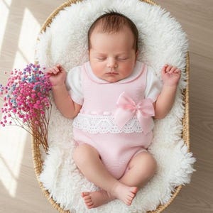 May include: A newborn baby wearing a pink romper with a white lace trim and a large bow. The baby is lying in a woven basket on a fluffy white blanket, with a bouquet of colorful flowers nearby.