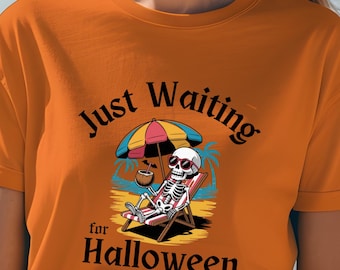 May include: An orange t-shirt featuring the text "Just Waiting for Halloween." The graphic depicts a skeleton lounging on a beach chair beneath a colourful umbrella, holding a coconut drink. The design uses black, white, and shades of blue and yellow.