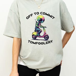 May include: A light green t-shirt with a graphic of an alien riding a scooter. The text "Off to Commit Tomfoolery" is printed on the shirt.