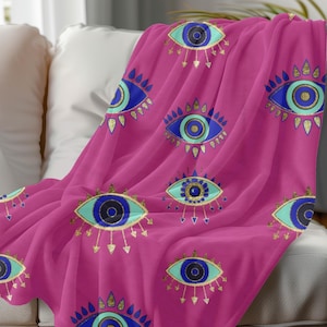 May include: Pink fleece throw blanket with a repeating pattern of blue and gold evil eyes. The eyes have a turquoise outline and gold accents.