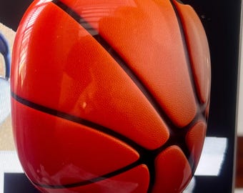May include: A basketball-themed decorative object, featuring a glossy orange and black design. The object is spherical, with black lines mimicking a basketball's panels. The surface has a textured appearance, and a silver metal loop is attached at the top.