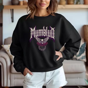 May include: Black sweatshirt featuring the word "Mumblud" in a gothic font above a skull with wings and floral accents. The design is in shades of purple and white. The sweatshirt is a crew neck style.