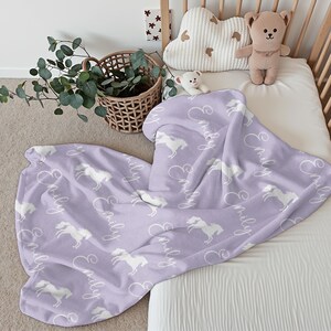 Pu&ograve; includere: Una coperta per neonati lilla con un motivo di cavalli bianchi e il nome "Emily" in corsivo. La coperta &egrave; appoggiata su una culla, con un orsacchiotto e un cuscino a forma di nuvola.